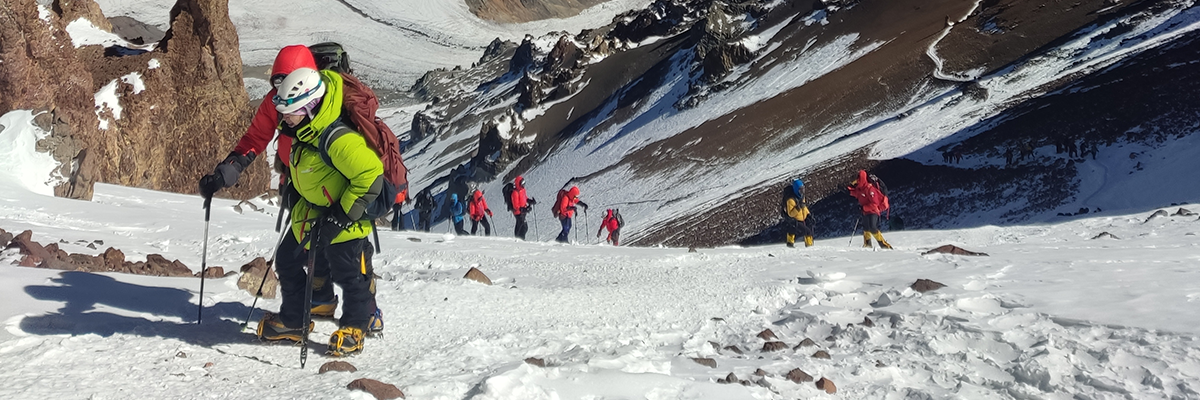 Montañistas en fila ascendiendo por una ladera nevada del Aconcagua, con el cielo azul de fondo.